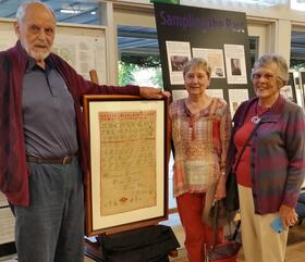 Trevor Shumack, Margaret Shumack's great nephew, Sandra Whichelo who donated the sampler to the Centre and Sandra Shumack, Trevor's wife showcasing the sampler at St. Ninian's Hall in Lyneham.